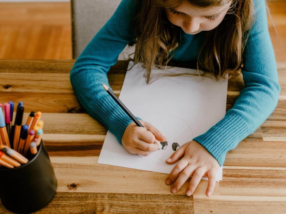 A young girl drawing on paper with a pencil at a wooden table.